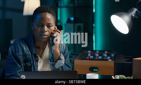Femme recevant l'appel téléphonique d'un ami tout en travaillant de la maison sur ordinateur portable dans le bureau personnel. Freelance parlant avec mate en ligne à l'aide du téléphone portable tout en résolvant des tâches, caméra A. Banque D'Images