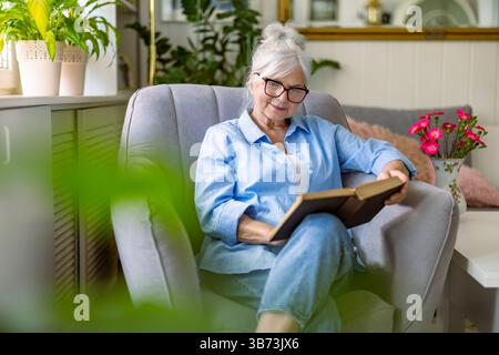 Femme mature lisant un livre dans un fauteuil confortable à la maison Banque D'Images