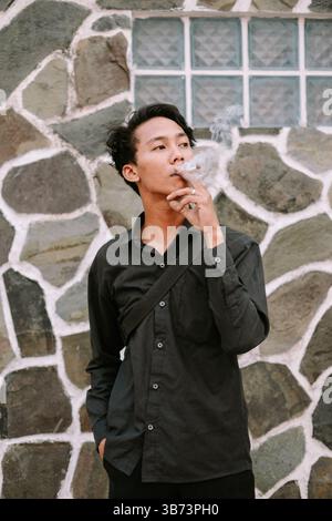 Un jeune homme fume une cigarette dans un cadre décontracté. Il respire des vibrations pensives contre un mur texturé. Banque D'Images