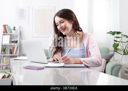 Jeune femme heureuse à la maison, assise à un bureau, utilisant son ordinateur portable et prenant des notes avec un stylo dans un cahier. Concept de travail à distance positif, lear en ligne Banque D'Images