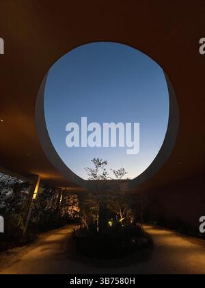 Une ouverture architecturale circulaire encadre le ciel crépusculaire sur un jardin de cour minimaliste, mêlant nature et design dans un espace moderne serein. Banque D'Images