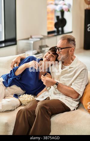 Couple d'âge moyen embrasse joyeusement sur leur canapé tout en dégustant du pop-corn ensemble. Banque D'Images