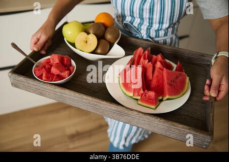 Un gros plan d'une personne tenant un plateau en bois avec des fruits frais, y compris des tranches de pastèque vibrantes et une variété de fruits mûrs dans un bol, favorisant h. Banque D'Images
