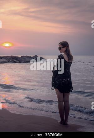 Une femme en vêtements de plage noirs et lunettes de soleil rencontre le coucher du soleil sur la côte de Kaolak, pose - demi-retournée. Belle vue sur le bord de mer rocheux. Banque D'Images