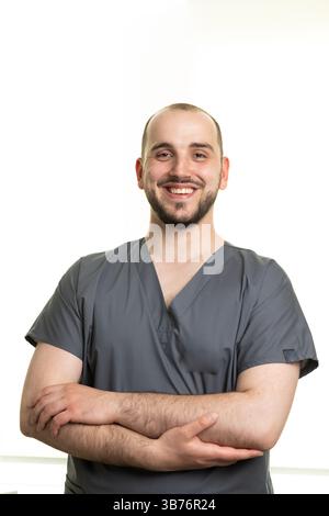 Un travailleur de la santé masculin joyeux portant des gommages gris se tient confiant avec les bras croisés, affichant un sourire amical dans un style minimaliste bien éclairé Banque D'Images