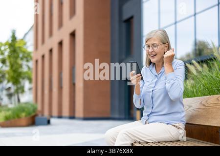 Une femme âgée excitée portant des lunettes regarde son téléphone, célébrant les bonnes nouvelles tout en étant assise dehors. Elle est heureuse. Banque D'Images