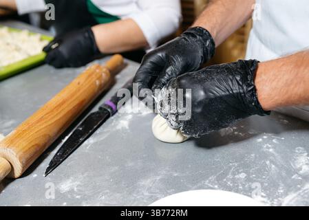 Gros plan des mains pliant la pâte pour faire des boulettes traditionnelles, avec rouleau à pâtisserie et couteau sur le comptoir fariné dans la cuisine. Banque D'Images