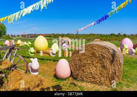Cadre de Pâques dynamique avec de grands œufs de couleur pastel et des lapins décoratifs dispersés autour d'un champ herbeux avec des balles de foin et des banderoles. Banque D'Images