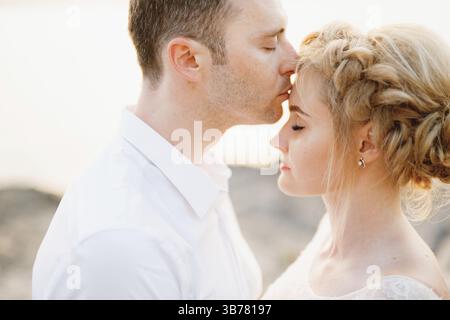 Le marié embrasse doucement la mariée sur le front sur les rochers au bord de la mer, gros plan. Banque D'Images