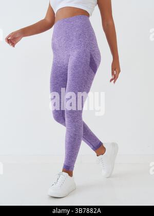 Femme athlétique en leggings violets taille haute et baskets blanches, top blanc court. Mouvement de marche capturé à mi-pas avec accent sur l'élégance Banque D'Images