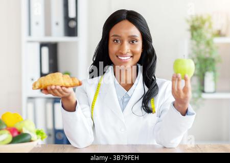 Faire des choix alimentaires sains. Belle nutritionniste féminine tenant croissant et pomme à table dans le bureau médical Banque D'Images