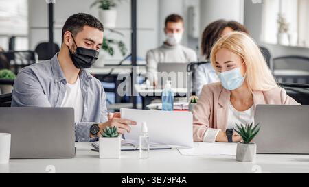 Distance sociale et travail avec des documents dans un bureau moderne. Homme et femme milléniaux avec des papiers dans le lieu de travail avec des ordinateurs portables et bouteille de discours antiseptique Banque D'Images