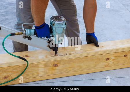 Le travailleur pousse les clous dans la poutre en bois en utilisant le pistolet à clous à air sur le site de construction sous la journée de travaux Banque D'Images