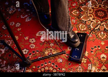 Plan à angle élevé de pied de femme portant des bottes appuyant sur le bouton sur le pédalier de guitare contrôlant les effets audio, tout en jouant de la musique rock en studio avec tapis rétro Banque D'Images