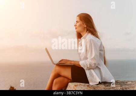 Femme Laptop Sunset - Une femme est assise sur une falaise avec un ordinateur portable, regardant le coucher du soleil sur l'océan. Banque D'Images