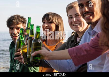 Gros plan de divers groupes d'amis acclamant avec des bouteilles de bière lors d'une fête estivale au bord de la mer. Banque D'Images