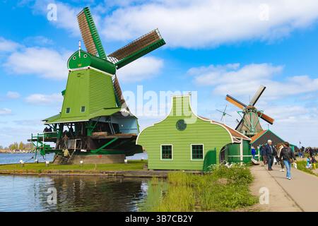 Le grand moulin à vent vert de la scierie 'de Gekroonde Poelenburg' et un petit bâtiment se tiennent près d'un canal à Zaanse Schans, pays-Bas, avec des gens sur un chemin. Banque D'Images