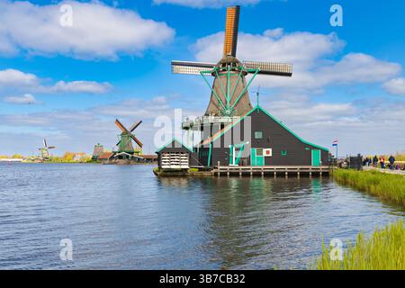Le moulin à vent de peinture 'de Kat' se tient au bord de l'eau à Zaanse Schans, aux pays-Bas, avec d'autres moulins à vent au loin, un chemin, des gens et un ciel nuageux. Banque D'Images