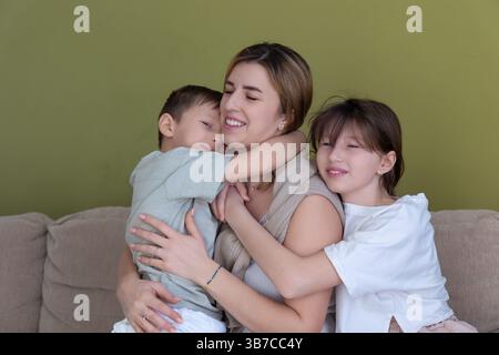 Mère aimante embrassant ses enfants à la maison. Une mère heureuse est assise sur un canapé, serrant chaleureusement ses deux enfants comme ils sourient et embrassent, partageant un Banque D'Images