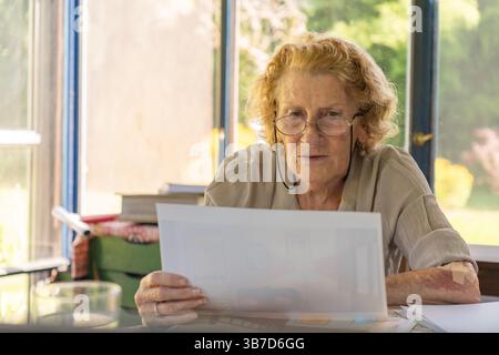 Une femme âgée avec des lunettes est assise à une table dans une pièce ensoleillée, examinant attentivement un document qu'elle tient dans ses mains, peut-être travaillant avec lui Banque D'Images