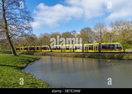 Un long tramway jaune longe un canal par une journée ensoleillée dans une zone verdoyante et arborée. 9 avril 2025. Banque D'Images