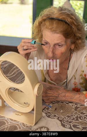 Femme âgée se maquillant devant un miroir grossissant, assise à une table avec une nappe de dentelle, démontrant une routine de beauté pour mature in Banque D'Images