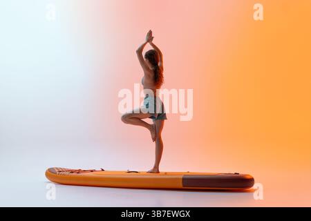 Femme sportive effectue une pose d'arbre debout à bord avec les bras levés au-dessus de la tête en équilibre, brillant doucement sous les lumières du studio. Banque D'Images
