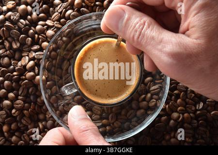 Détail des mains remuant du café fraîchement infusé dans une tasse en verre et une soucoupe avec une cuillère en métal sur un tas de grains de café. Vue de dessus. Banque D'Images