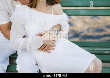 Un homme et une femme en vêtements blancs sont assis sur un banc vert, un homme embrasse doucement une femme par derrière, gros plan. Banque D'Images