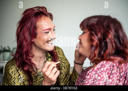 Personne non binaire souriante avec de longs cheveux roux et le maquillage applique le maquillage à leur ami, célébrant l'acceptation de soi, la positivité du corps, et le sexe F. Banque D'Images