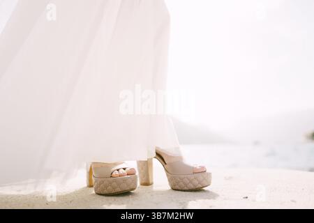 Jambes de mariée dans une robe blanche et chaussures beiges à talons hauts sur la jetée. Gros plan. Banque D'Images
