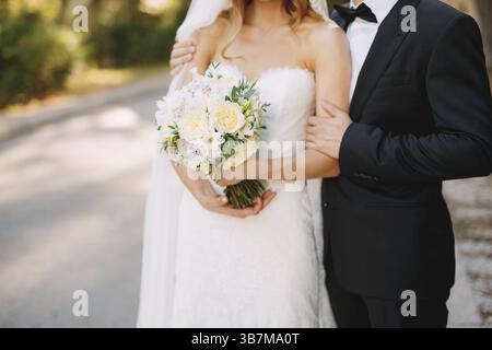 Le marié embrasse les épaules de la mariée, qui tient un bouquet de mariage avec des fleurs blanches, gros plan . Banque D'Images
