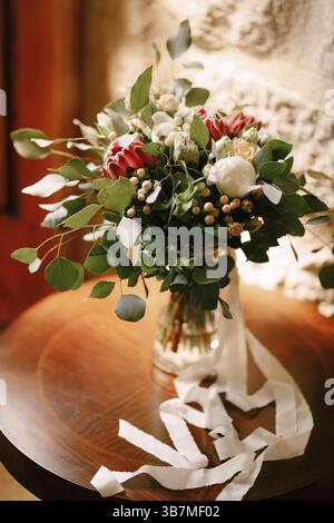 Bouquet de mariée de pivoines blanches, roses, protéa rose, baie de neige, branches d'eucalyptus et rubans blancs sur la table ronde en bois. Banque D'Images
