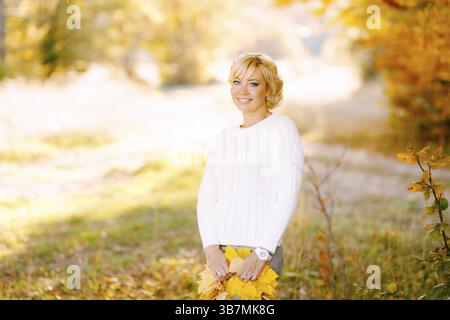 Demi-portrait de femme souriante debout avec un bouquet de feuilles jaunes dans la forêt d'automne. Banque D'Images
