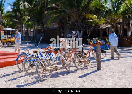 Holbox, Mexique - 9 février 2025 : des vélos de plage et un petit stand de fruits sont assis sous des palmiers oscillants dans une rue sablonneuse à Holbox, au Mexique. Banque D'Images