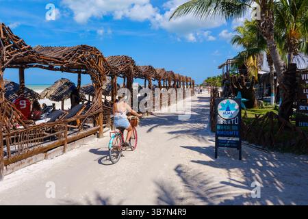 Holbox Island, Mexique – 10 février 2025 : une femme fait du vélo de croisière le long d'un chemin sablonneux bordé de cabanes de plage de bois flotté et de palmiers, passant par b Banque D'Images