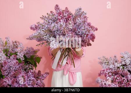 Femme cachant le visage derrière un grand bouquet de lilas florissants sur fond rose, concept floral de printemps. Banque D'Images