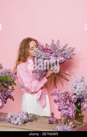 Femme joyeuse appréciant le bouquet de lilas parfumé dans un studio floral printanier avec un fond rose pastel doux. Banque D'Images