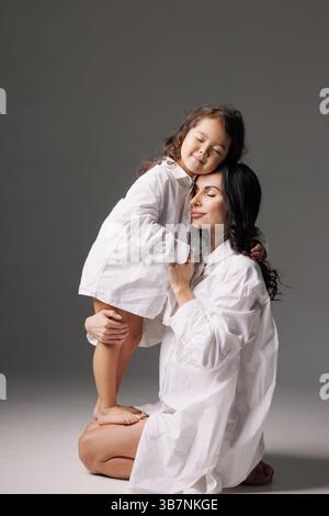 Mère aimante embrassant sa fille pieds nus dans des chemises blanches surdimensionnées, les yeux fermés, moment familial tendre. Banque D'Images