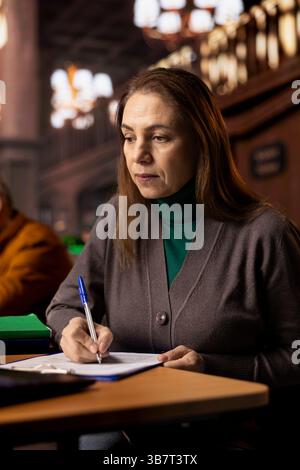Étudiant ciblé prenant des notes d'un webinaire et examinant des fichiers archivés dans une salle de travail de bibliothèque, montrant le dévouement de l'éducation mature et du développement des connaissances. Travailler sur sa thèse. Banque D'Images