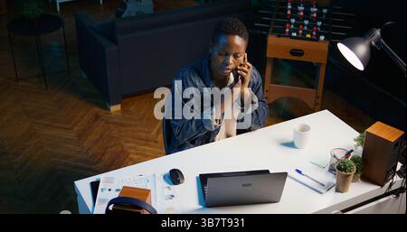 Télétravailleur afro-américain recevant un appel téléphonique d'un collègue de travail tout en travaillant à domicile sur un ordinateur portable. Freelance parlant avec un collègue à l'aide d'un smartphone tout en résolvant des tâches, caméra B. Banque D'Images