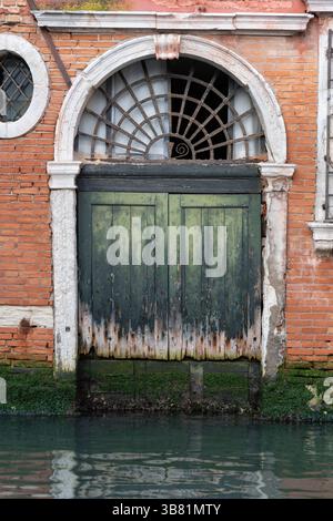 Vieille porte en bois vieilli avec treillis arqué et cadre en pierre au canal dans la ville de Venise, Italie. Banque D'Images