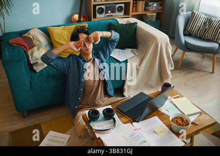 Jeune homme prenant une pause du travail tout en étant assis sur un canapé confortable dans un salon rempli de papiers, livres et ordinateur portable. Une installation de bureau à domicile décontractée avec esprit Banque D'Images