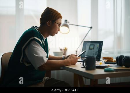 Jeune homme assis au bureau à l'aide d'un smartphone tout en travaillant sur un ordinateur portable. Il porte des lunettes et un bonnet, et il y a divers articles sur le bureau, y compris bo Banque D'Images