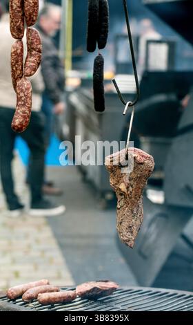 Viandes salées traditionnelles accrochées à des crochets au marché de rue. Saucisses fumées et viande vieillie affichées à l'étal de boucherie avec grill au premier plan. Banque D'Images