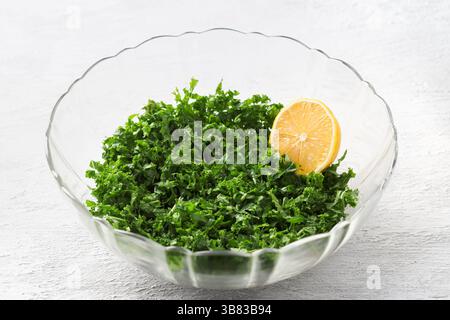 Feuilles de chou frisé hachées écrasées avec du jus de citron, un demi-citron dans un saladier transparent, préparant une salade maison saine sur un dos gris clair Banque D'Images