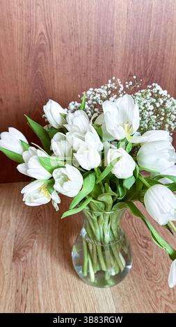 Élégant bouquet de tulipes blanches dans un vase en verre avec le souffle de bébé sur une surface en bois capturant l'essence du décor floral minimaliste. Banque D'Images