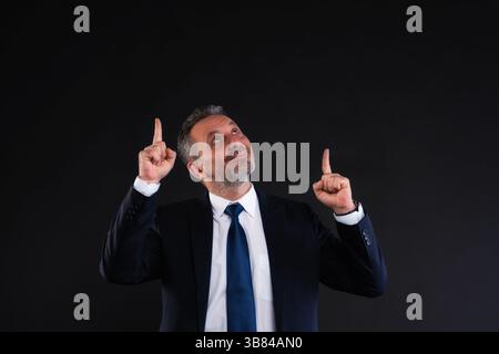 Homme d'affaires confiant en costume souriant et pointant vers le haut avec les deux index, suggérant le succès, le progrès ou l'opportunité. Studio tourné en noir Banque D'Images