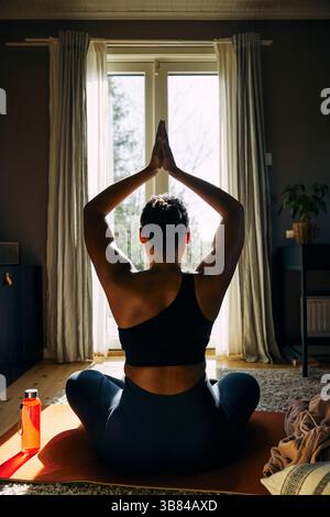 Vue arrière de la femme avec faire du yoga avec la main serrée tout en étant assis à la maison Banque D'Images