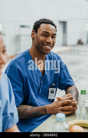 Expert médical masculin souriant assis les mains serrées dans la cafétéria de l'hôpital Banque D'Images